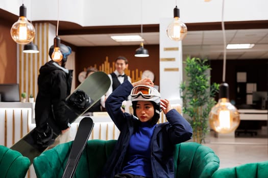 Ski mountain resort lounge area filled with young customers in winter jackets and helmets. Asian woman on couch in hotel lobby adjusting ski goggles preparing for fun wintersport activity.