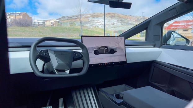 A driver perspective inside the Tesla Cybertruck, highlighting its spacious, minimalist interior with a large touchscreen display and futuristic steering wheel, set against a backdrop of a scenic landscape.