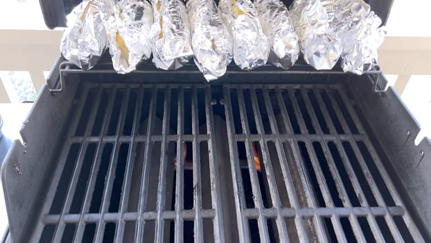 Several ears of corn wrapped in aluminum foil are lined up on a barbecue grill, slowly roasting to perfection, showcasing a popular and delicious method of cooking this classic side dish.