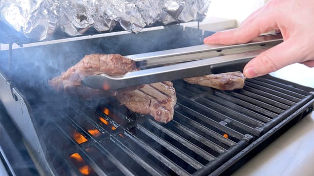 A person uses tongs to flip juicy steaks on a grill, with flames licking the meat, captured in a moment of intense heat and sizzle, showcasing a classic outdoor cooking experience.