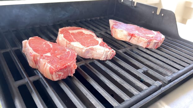 This image showcases the art of grilling, featuring three thick steaks cooking on a barbecue grill, with a row of foil-wrapped corn on the cob above, capturing a typical scene of a hearty outdoor meal preparation.