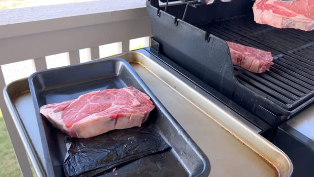 This image showcases the art of grilling, featuring three thick steaks cooking on a barbecue grill, with a row of foil-wrapped corn on the cob above, capturing a typical scene of a hearty outdoor meal preparation.