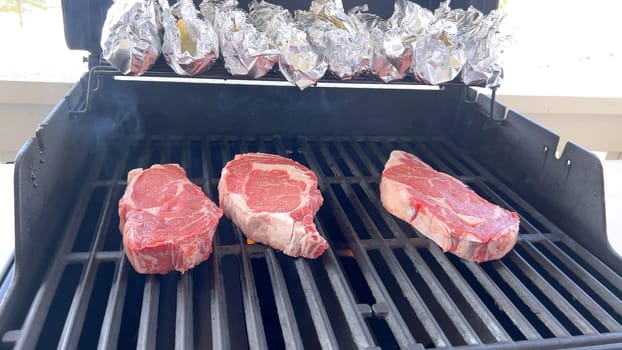 This image showcases the art of grilling, featuring three thick steaks cooking on a barbecue grill, with a row of foil-wrapped corn on the cob above, capturing a typical scene of a hearty outdoor meal preparation.