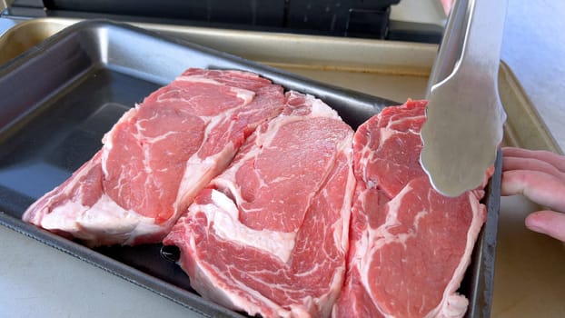 This image showcases the art of grilling, featuring three thick steaks cooking on a barbecue grill, with a row of foil-wrapped corn on the cob above, capturing a typical scene of a hearty outdoor meal preparation.