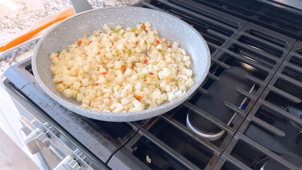 A frying pan on a gas stove sizzles with diced potatoes and colorful bits of red and green peppers, showcasing a delicious and simple home-cooked side dish in the making.