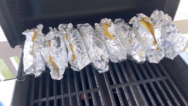 Several ears of corn wrapped in aluminum foil are lined up on a barbecue grill, slowly roasting to perfection, showcasing a popular and delicious method of cooking this classic side dish.