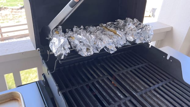 Several ears of corn wrapped in aluminum foil are lined up on a barbecue grill, slowly roasting to perfection, showcasing a popular and delicious method of cooking this classic side dish.