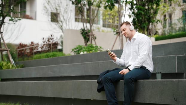 Smart businessman wear headphone to listen relaxed music while take off suit and move to music at green city. Side view of happy project manager with headset dancing movement with lively song. Urbane.