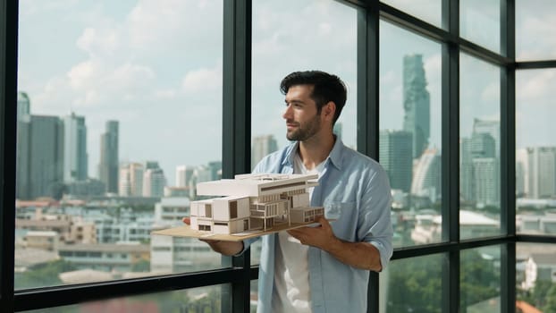 Skilled smiling caucasian engineer turns house model left and right to check mistake point. Professional manager inspect at architectural model while standing near window. Design concept. Tracery