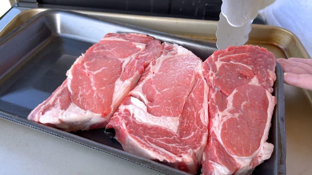 This image showcases the art of grilling, featuring three thick steaks cooking on a barbecue grill, with a row of foil-wrapped corn on the cob above, capturing a typical scene of a hearty outdoor meal preparation.