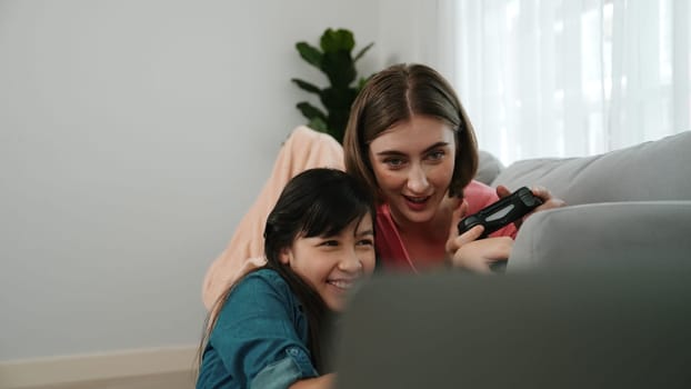 Happy girl playing game and sitting while mom lie on sofa by using laptop screen. Caucasian parent and attractive girl spend time together while holding joystick and focus on winning games. Pedagogy.