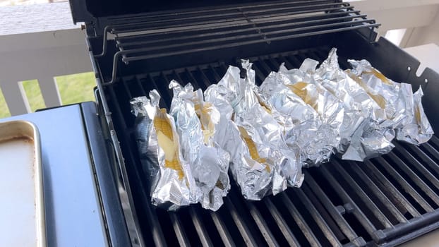 Several ears of corn wrapped in aluminum foil are lined up on a barbecue grill, slowly roasting to perfection, showcasing a popular and delicious method of cooking this classic side dish.