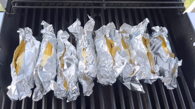 Several ears of corn wrapped in aluminum foil are lined up on a barbecue grill, slowly roasting to perfection, showcasing a popular and delicious method of cooking this classic side dish.