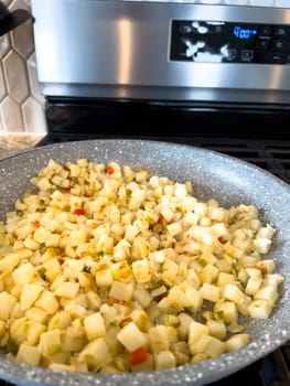 A frying pan on a gas stove sizzles with diced potatoes and colorful bits of red and green peppers, showcasing a delicious and simple home-cooked side dish in the making.