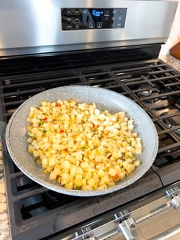 A frying pan on a gas stove sizzles with diced potatoes and colorful bits of red and green peppers, showcasing a delicious and simple home-cooked side dish in the making.