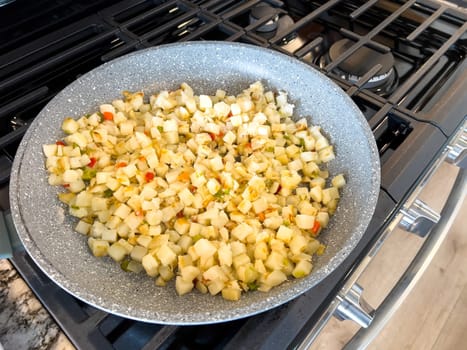 A frying pan on a gas stove sizzles with diced potatoes and colorful bits of red and green peppers, showcasing a delicious and simple home-cooked side dish in the making.