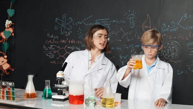Caucasian teacher helps student mixed liquid in science laboratory. Professional instructor support smart boy doing experiment in STEM science class at blackboard written chemical theory. Erudition.