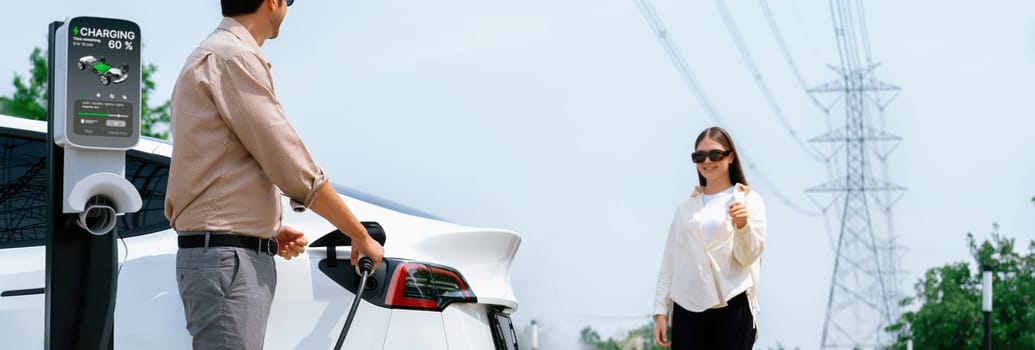 Young couple recharge EV car battery at charging station connected to power grid tower electrical industrial facility as electrical industry for eco friendly vehicle utilization. Panorama Expedient