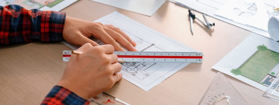 Closeup of architect engineer hand using ruler to mature and draw a blueprint on meeting table with wooden block, pencil and blueprint scatter around at architectural modern office. Delineation.