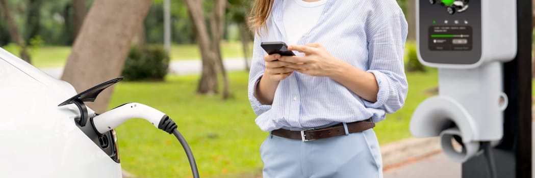 Young woman using smartphone online banking application to pay for electric car battery charging from EV charging station during vacation road trip at national park or summer forest. Panorama Exalt