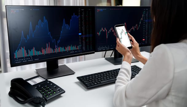 Smiling young Asian businesswoman with happy face looking on smartphone in profitable of exchange stock market graph at modern office. Concept of investing high profit in wealth security. Stratagem.