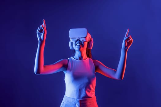 Smart female stand with surrounded by cyberpunk neon light wear VR headset connecting metaverse, futuristic cyberspace community technology. Woman using finger pointing virtual object. Hallucination.