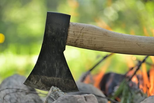 Closeup view of an axe with a wooden handle, embedded in a log, with the warm glow of a campfire in the background, survival or historical recreation, leisure activities and relaxation around a fire