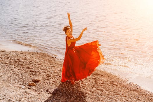 Woman red dress sea. Female dancer in a long red dress posing on a beach with rocks on sunny day. Girl on the nature on blue sky background