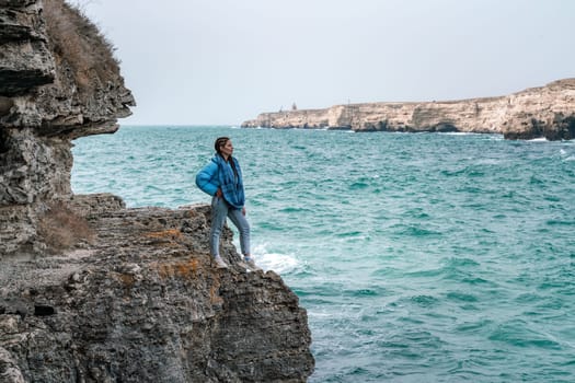 A woman in a blue jacket stands on a rock above a cliff above the sea and looks at the raging ocean. Girl traveler rests, thinks, dreams, enjoys nature. Peace and calm landscape, windy weather