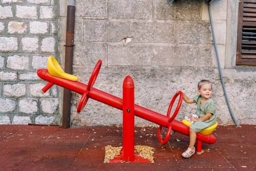 Little girl sits on a swing-balancer near an old house and looks away. High quality photo