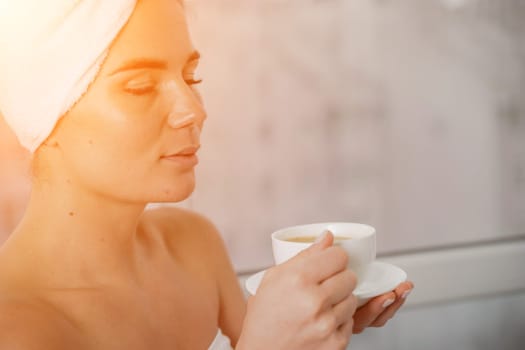 Young serene relaxed woman in spa bath towel drinking hot beverage tea coffee after taking shower bath at home. Beauty treatment, hydration concept