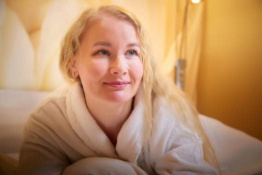 A blonde girl in white terry bathrobe in a hotel room after spa treatments on a trip or travel. Weekend and vacation body care