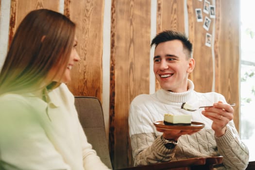 A cheerful young man, dressed in a white sweater, shares a delightful moment with his friend over a slice of cheesecake at a warm, inviting cafe. Their joyful interaction suggests a comfortable friendship as they savor the dessert together, surrounded by a wooden interior that adds to the cozy ambiance.