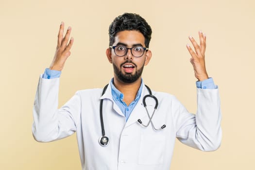 Oh my God, Wow. Excited amazed winner Indian young doctor cardiologist man surprise looking at camera with big eyes, shocked by victory celebrating success. Apothecary pharmacy guy on beige background