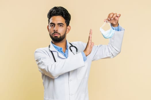 Happy Indian young doctor cardiologist man removing, taking off medical protective mask. Flu treatment cure. Ending of pandemic quarantine. Arabian apothecary pharmacy guy isolated on beige background