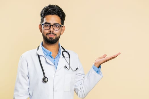 Indian young doctor cardiologist man show thumbs up and pointing right empty place, advertising area for commercial discount, copy space. Arabian apothecary pharmacy guy isolated on beige background