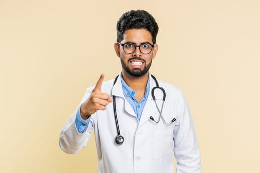 Indian young doctor cardiologist man shakes finger and saying no, be careful, advice to avoid danger mistake, disapproval sign, disagree, dislike. Arabian apothecary pharmacy guy on beige background