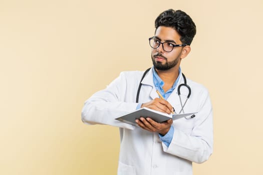 Thoughtful Indian young doctor cardiologist man making notes, writing down thoughts with pen into notepad notebook diary, to do list, good idea. Arabian apothecary pharmacy guy on beige background