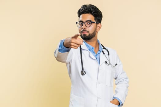 Hey you. Happy strict Indian young doctor cardiologist man pointing forward to camera, choosing, indicating to you, promotion. Arabian apothecary pharmacy smiling guy isolated on beige background