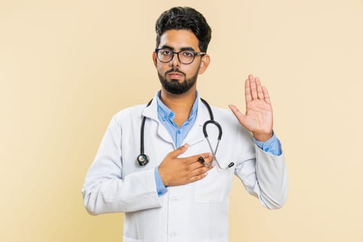 I swear to be honest. Sincere responsible Indian young doctor cardiologist man raising hand to take oath, promising to tell truth, keeping hand on chest. Apothecary pharmacy guy on beige background