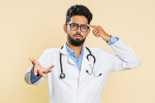 You are crazy, out of mind. Confused Indian young doctor cardiologist man pointing at camera and showing stupid, bullying, asking why fail bad plan. Arabian apothecary guy isolated on beige background