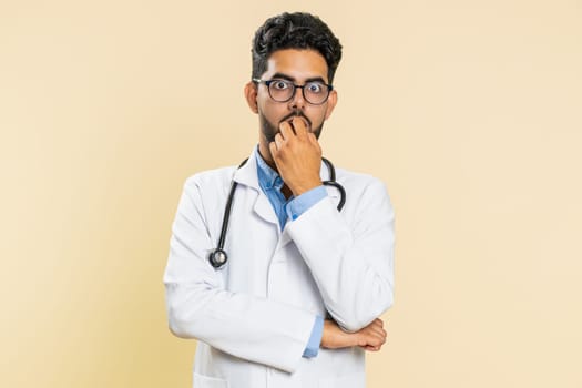 Upset scared frightened Indian doctor cardiologist man biting nails, feeling worried nervous about serious troubles, stress, anxiety disorder, panic attack, guilty. Arabian guy on beige background