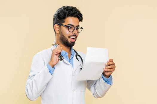 Indian young doctor cardiologist man open envelope take out letter reads it feel happy. Career growth advance promotion, long-awaited invitation great news. Arabian apothecary guy on beige background