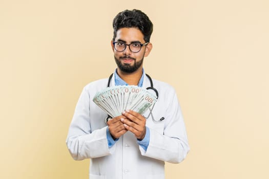 Rich happy Indian doctor cardiologist man waving money dollar cash banknotes bills like a fan, success business career, big income wealth. Arabian apothecary pharmacy guy isolated on beige background