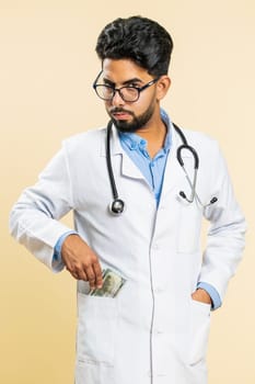 Indian speaker doctor cardiologist man receiving bribe from patient, taking money from client, hiding cash into pocket. Illegal corruption in medicine. Arabian apothecary guy on beige background