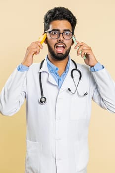 Irritated nervous stressed tired Indian doctor cardiologist man talking screaming on two mobile phones have conversation conflict quarrel. Arabian apothecary pharmacy guy on beige background. Vertical