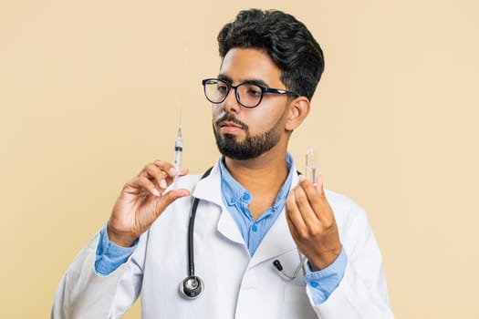 Indian young doctor cardiologist man holds syringe needle and ampoule tube with medical vaccine medicine treatment injection ready to use. Immunization. Arab scientist guy isolated on beige background