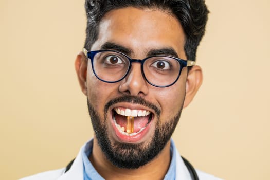 Smiling Indian doctor man puts of tablet pill vitamin D or omega-3 yellow capsule into mouth, recommends drugs immunization cure treatment. Health care. Apothecary pharmacist on beige background