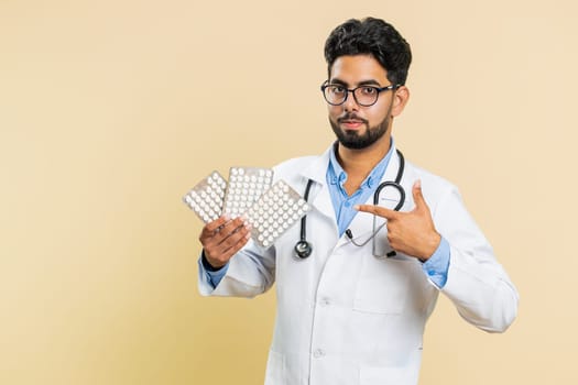 Indian doctor cardiologist man shows lot of tablet pills in blisters, recommends drugs vitamin antibiotic cure treatment. Health care. Arabian apothecary pharmacist guy isolated on beige background