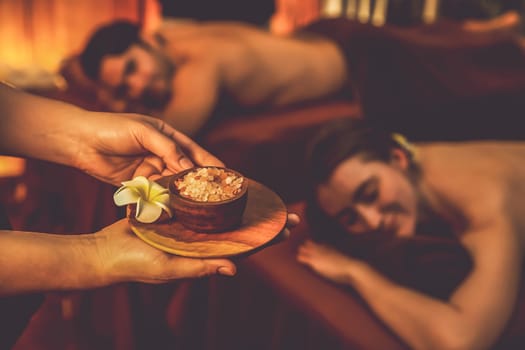 Masseur holding spa salt while couple customer having exfoliation treatment in luxury spa salon with warmth candle light ambient. Salt scrub beauty treatment in Health spa body scrub. Quiescent